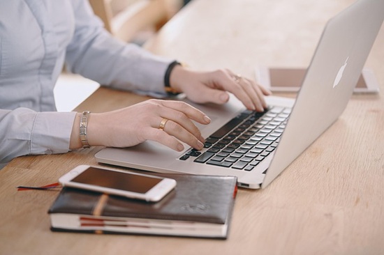 On a table sit an open laptop, a leather-bound book with a smartphone on top, and a tablet in the background. A woman's hands are typing on the laptop's keyboard.