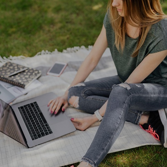 A white blanket is spread on grass. It holds an open laptop, notebook and pen, white smartphone. A young woman with long hair, wearing a tee-shirt and stylishly-ripped jeans, is sitting on the blanket studying the laptop screen. Her hand is positioned to use the mouse touchpad.