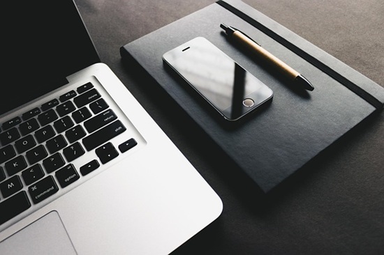 Image shows an open laptop and a smart phone and pen resting on a closed hardback journal.