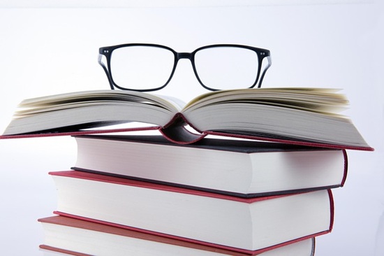 A stack of hardcover books, with the top book lying open and a pair of glasses on top.