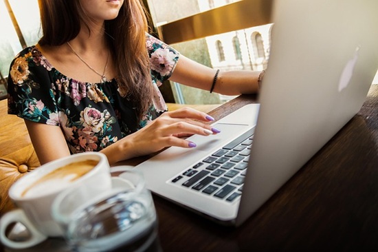 A woman in a flowered top works at a table with a laptop. A cup of coffee and glass of water are in the foreground.